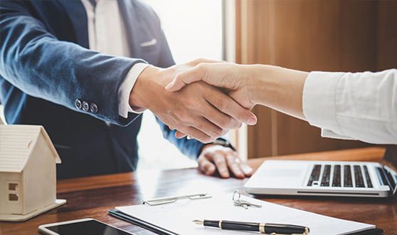 Two people shaking hands over a wooden desk with a laptop, documents, and a small house model in the foreground.