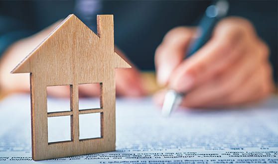 A small wooden house model sits on a document, while a person in the background signs paperwork with a pen.