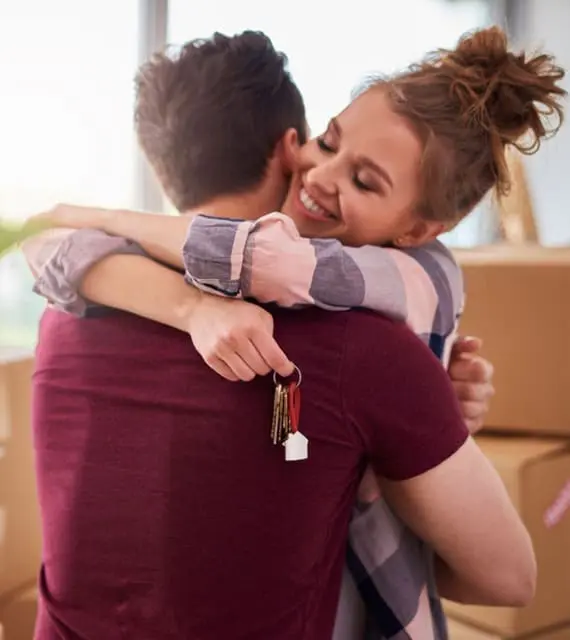 A woman with a bun hugs a man in a red shirt while holding a set of keys. Both are standing in a room with moving boxes.