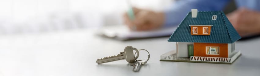 A pair of keys lies on a white surface next to a small model house, with a person writing on a document in the background.