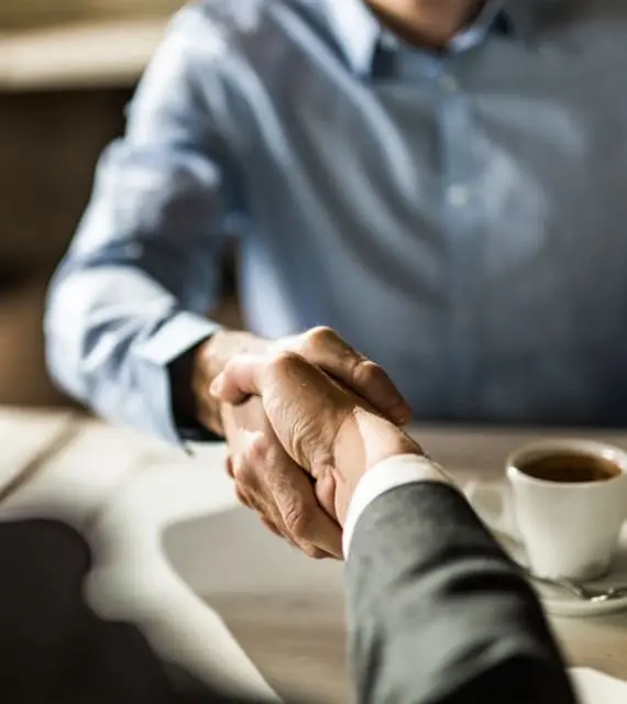 Two people shaking hands over a table with a cup of coffee in the background. Only their hands and part of their torsos are visible.