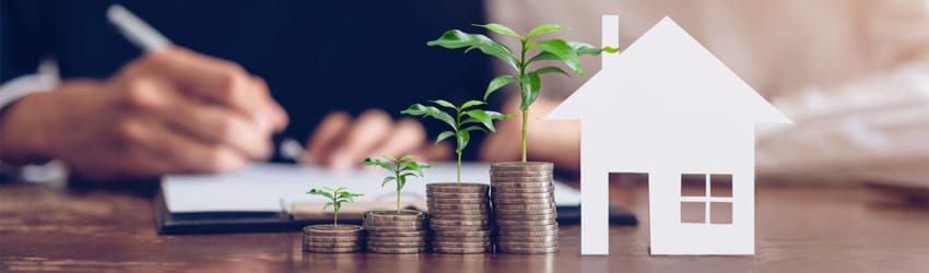 A person writes in a notebook, while stacks of coins with growing plants and a paper house model are on the table in the foreground.