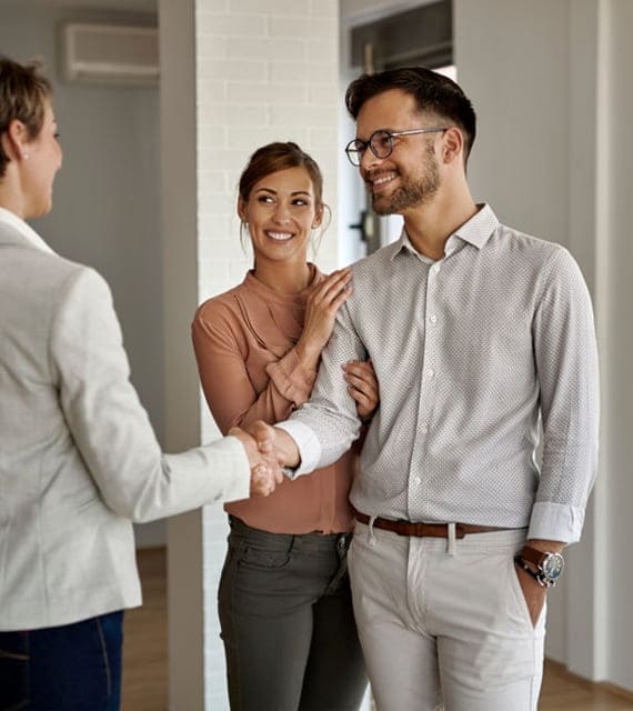 A couple, with the woman wearing a pink blouse and the man in a light-colored shirt, smiles while the man shakes hands with another person in a light-colored blazer.